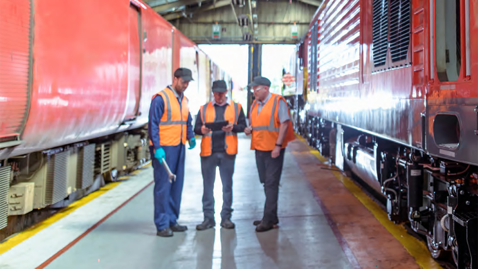Three workers wearing orange vests and safety equipment stand in between two parked trains